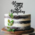 A tiered cake with a light cream and dark chocolate exterior, topped with a black acrylic cake topper that reads "Happy 25th Birthday." The cake is decorated with green leaves along the side and sits on a round wooden cake stand. The background is softly blurred.