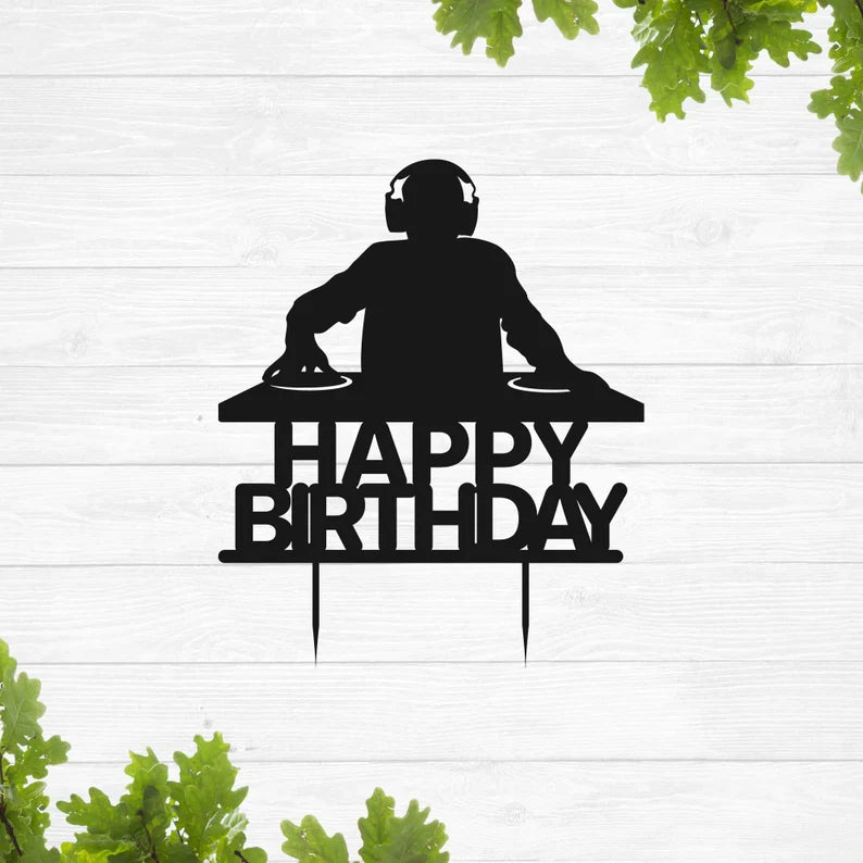 A silhouette of a DJ wearing headphones is shown in front of a turntable setup. Below the DJ, the text "HAPPY BIRTHDAY" is displayed in bold, stylized letters. The image has a light wooden background with green leaves framing the bottom. This cake topper design is suitable for birthday celebrations.