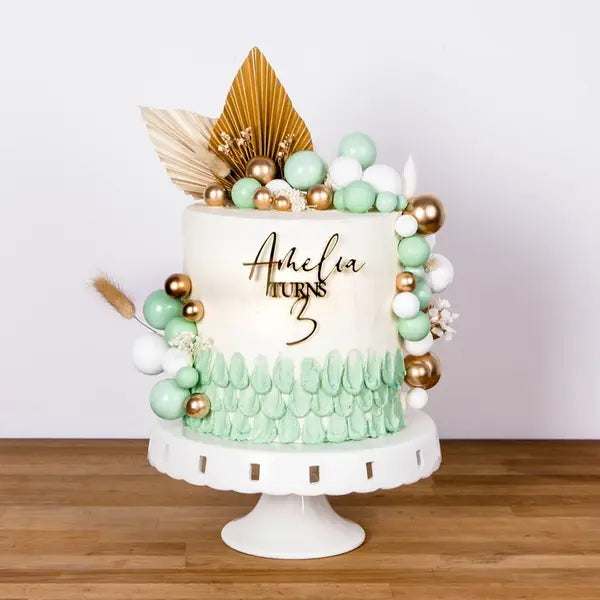 A decorated birthday cake sits on a white cake stand. The cake features a smooth white frosting with a mint green ruffled design at the base. It is adorned with colorful decorations, including green, gold, and white round spheres, and a cluster of leaves. The topper reads "Amelia TURNS 3" in a stylish font, highlighting the celebration. The background is neutral, emphasizing the cake&