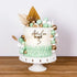 A decorated birthday cake sits on a white cake stand. The cake features a smooth white frosting with a mint green ruffled design at the base. It is adorned with colorful decorations, including green, gold, and white round spheres, and a cluster of leaves. The topper reads "Amelia TURNS 3" in a stylish font, highlighting the celebration. The background is neutral, emphasizing the cake&