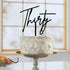A round cake with a textured white frosting sits on a decorative pedestal stand. Atop the cake is a black acrylic topper that reads "Thirty" in an elegant cursive font. The background features a window with soft lighting, and a plate with apples is visible in the foreground.