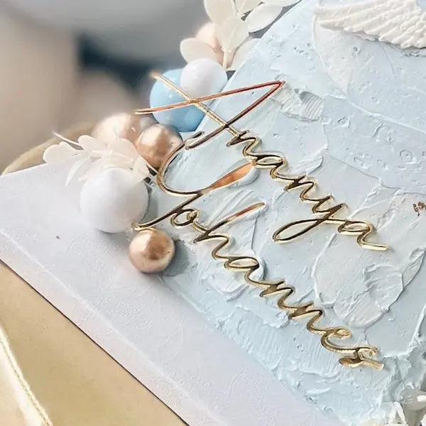The image features a beautifully decorated cake with a light blue textured surface. Atop the cake is a golden acrylic charm that reads "Ananya Johannes." Surrounding the charm are decorative elements, including shiny spheres in white, blue, and gold, along with delicate white floral accents. The overall presentation is elegant, suitable for a birthday celebration.
