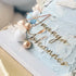 The image features a beautifully decorated cake with a light blue textured surface. Atop the cake is a golden acrylic charm that reads "Ananya Johannes." Surrounding the charm are decorative elements, including shiny spheres in white, blue, and gold, along with delicate white floral accents. The overall presentation is elegant, suitable for a birthday celebration.