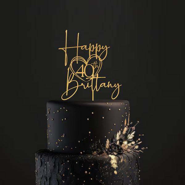 A black tiered cake adorned with a gold acrylic cake topper that reads "Happy 40th Brittany" styled with a heart. The cake features a textured surface with golden embellishments and decorative floral elements on one side, set against a dark background.