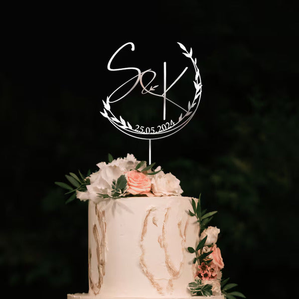 A personalized wedding cake topper featuring the initials "S&K" inside a circular design adorned with delicate leaves. Below the initials, the date "25.05.2024" is displayed. The topper is placed on a beautifully decorated wedding cake, which has a smooth white surface and subtle gold accents. The top of the cake is adorned with soft pink roses and greenery, adding a touch of elegance to the overall presentation. The background is dim, highlighting the cake and topper as the focal point.