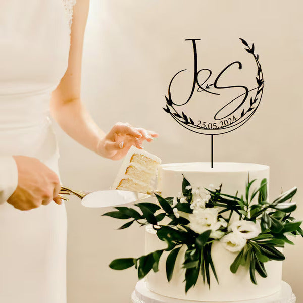 A bride, dressed in a white gown, is using a cake server to cut a slice from a multi-tiered wedding cake. The cake, adorned with fresh greenery and white flowers, features a personalised cake topper that reads "J&S" with a date "25.05.2024" encircled by a decorative leaf design.