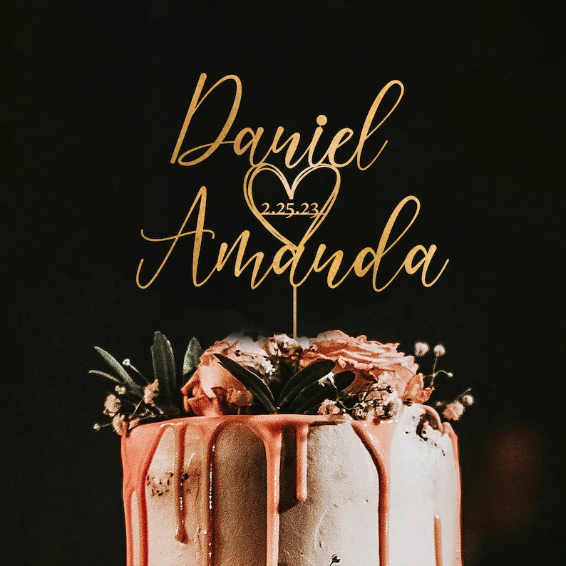 A wedding cake with a personalized acrylic cake topper featuring the names "Daniel" and "Amanda," the date "2.25.23," and a heart design. The cake is decorated with dripping pink frosting and adorned with flowers and greenery on top, set against a dark background.