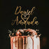A wedding cake with a personalized acrylic cake topper featuring the names "Daniel" and "Amanda," the date "2.25.23," and a heart design. The cake is decorated with dripping pink frosting and adorned with flowers and greenery on top, set against a dark background.