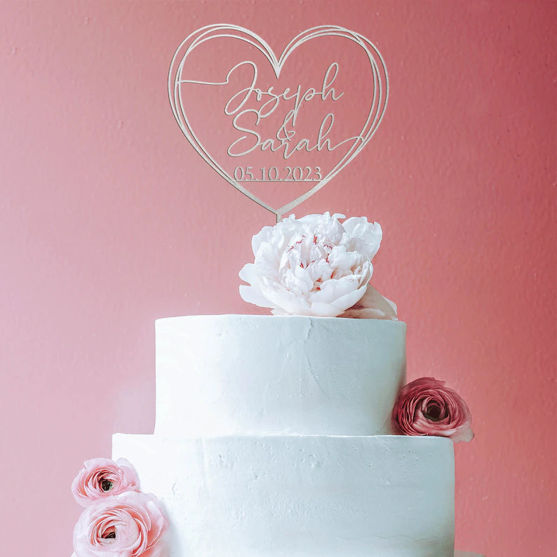 An elegant three-tier white wedding cake sits against a soft pink backdrop. Atop the cake is a personalized acrylic cake topper in a heart shape, featuring the names "Joseph" and "Sarah" along with the date "05.10.2023." The topper is clear and stylishly designed. Adorning the cake are delicate pink flowers, including a large peony at the top and smaller ranunculus flowers on the side, enhancing the romantic aesthetic.