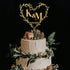 The image features a three-tiered wedding cake adorned with a heart-shaped acrylic cake topper. The topper is designed with the initials "K & M" and the date "02.03.2023," surrounded by delicate greenery. The cake itself has a textured white frosting and is decorated with light pink roses and various greenery elements on the top and around the base. The background is dark, providing contrast to the cake and the topper.