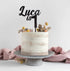 A round cake with a textured pale-colored frosting sits on a gray pedestal cake stand. Atop the cake, there is a black acrylic topper that reads "Luca is 1," elegantly styled in italic font. The cake is decorated with small, dried flowers and foliage in various colors, including yellow and green, which are arranged around the top. A soft pink cloth is draped in the foreground, adding a subtle, decorative touch to the presentation. The background is light and neutral, emphasizing the cake&