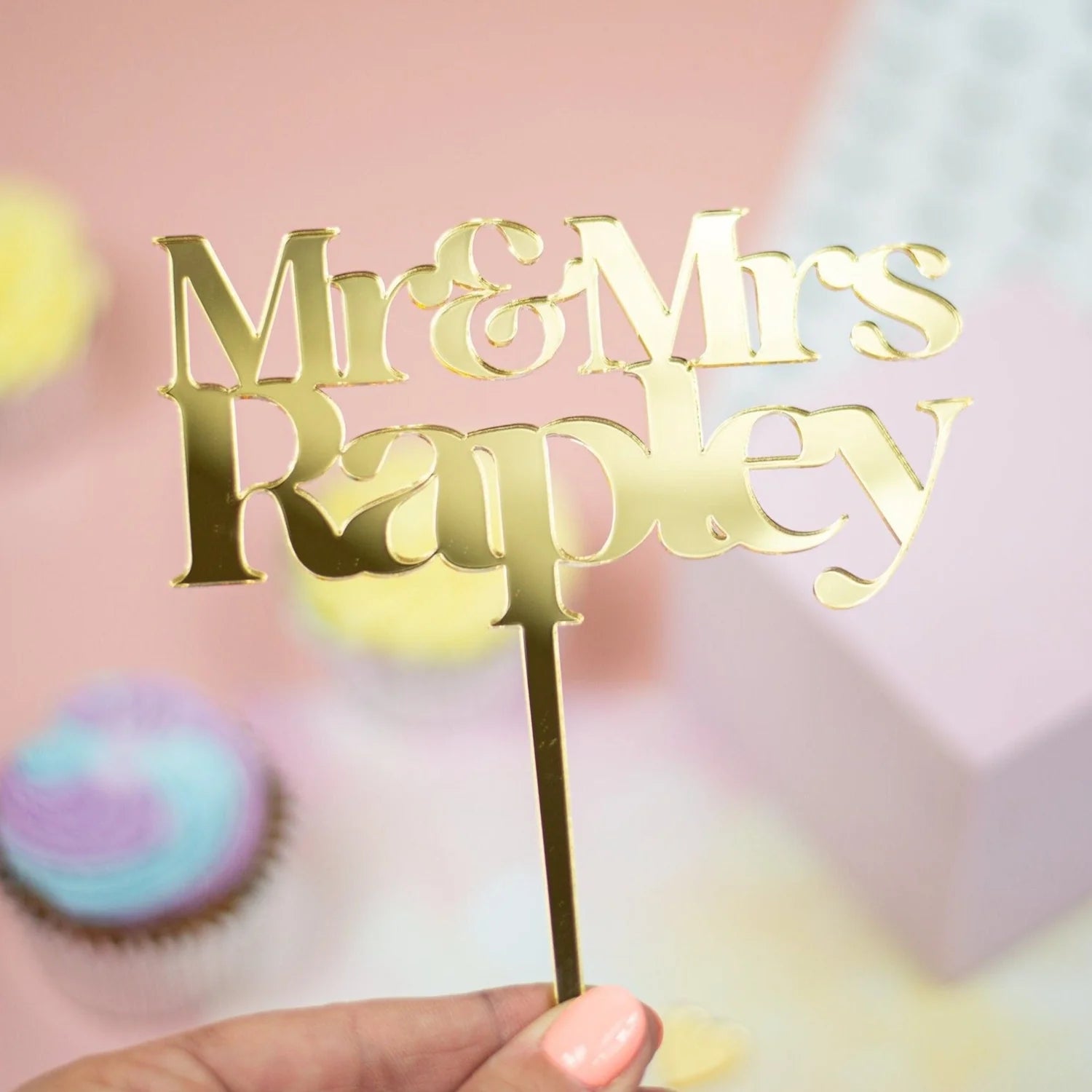 The image features a gold acrylic cake topper with the text "Mr & Mrs Rapley" in an elegant font. The topper is being held by a hand, and the background includes soft pastel colors, with cupcakes partially visible. The overall aesthetic is celebratory and suitable for a wedding or special occasion.