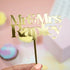 The image features a gold acrylic cake topper with the text "Mr & Mrs Rapley" in an elegant font. The topper is being held by a hand, and the background includes soft pastel colors, with cupcakes partially visible. The overall aesthetic is celebratory and suitable for a wedding or special occasion.