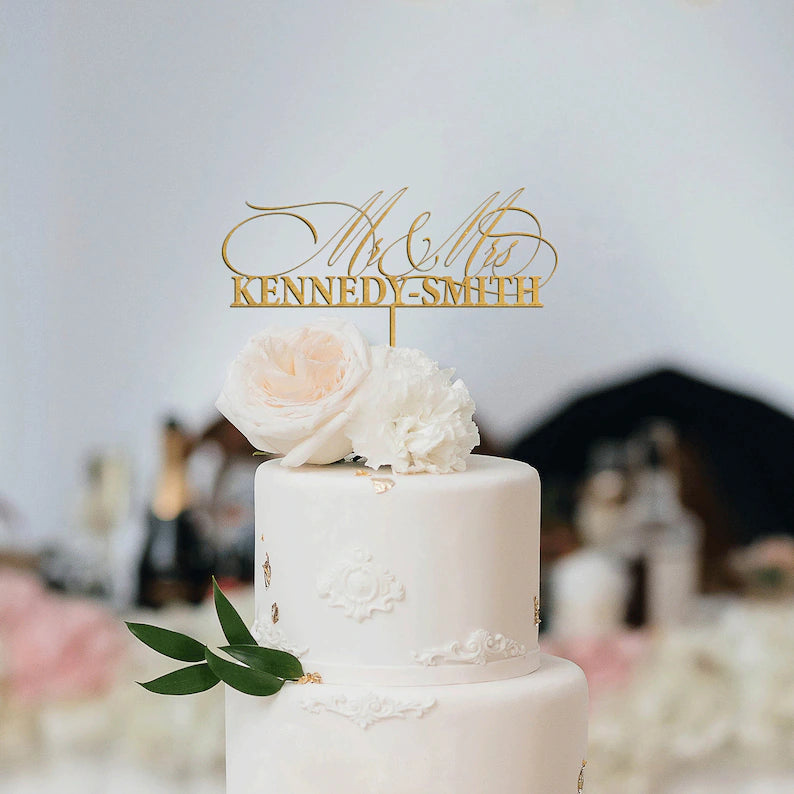 An elegant white wedding cake with two tiers decorated with floral elements. Atop the cake is a gold acrylic cake topper featuring the text ?Mr & Mrs Kennedy-Smith.? The cake is adorned with soft pink and white roses, along with a green leaf accent. The background suggests a celebratory setting with blurred decorations and items.
