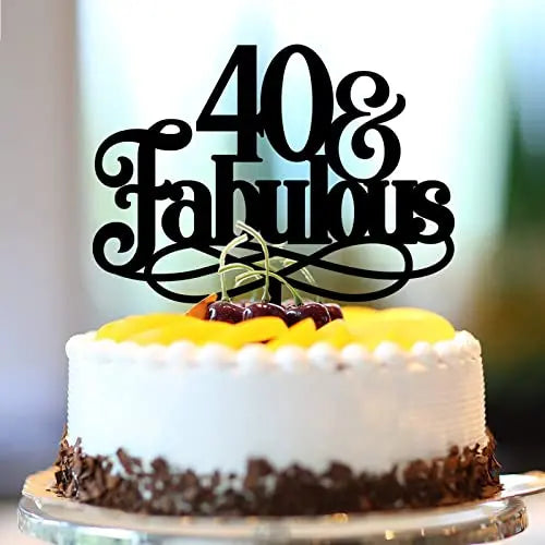 A round cake topped with a black decorative cake topper that reads "40 & Fabulous." The cake is frosted in white with a chocolate border and is garnished with yellow fruit on top, along with a few cherries. The background is softly blurred.