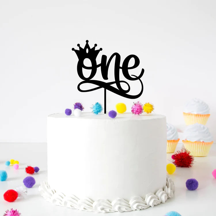 An acrylic cake topper featuring the word "one" in a stylish script font is placed on top of a white frosted cake, adorned with a small crown above the "o". The cake sits on a flat surface, surrounded by colorful pom-poms in various sizes, and nearby, there are pastel-colored cupcakes with white frosting. The background is white, creating a clean and festive presentation.