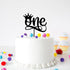 An acrylic cake topper featuring the word "one" in a stylish script font is placed on top of a white frosted cake, adorned with a small crown above the "o". The cake sits on a flat surface, surrounded by colorful pom-poms in various sizes, and nearby, there are pastel-colored cupcakes with white frosting. The background is white, creating a clean and festive presentation.