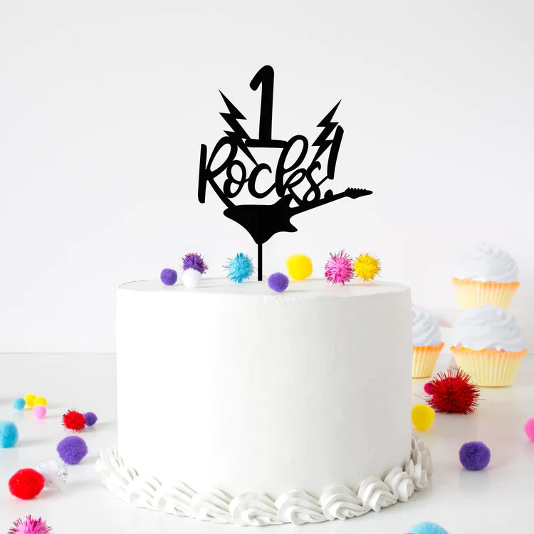 A round white cake topped with a black acrylic cake topper that reads "1 Rocks!" The topper features lightning bolt designs and a silhouette of a guitar. The cake is decorated with colorful pom-poms scattered around it, along with small cupcakes in the background. The setting has a bright, festive atmosphere.