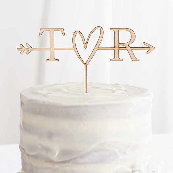 A personalized acrylic cake topper featuring the letters "T" and "R" on either side of a heart shape, with an arrow extending from the left side. The topper is displayed on a lightly frosted cake, showcasing a simple, elegant design. The backdrop is soft and neutral, enhancing the focus on the cake and topper.