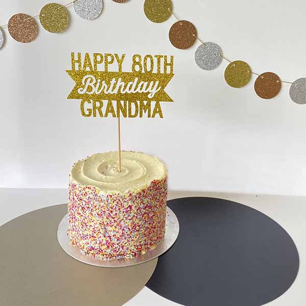 A round cake with a smooth vanilla frosting and colorful sprinkles on the sides sits on a silver cake board. Topped with a glittery gold cardstock cake topper that reads "HAPPY 80TH Birthday GRANDMA." In the background, there is a decorative garland made of circles in gold, silver, and brown. The table is covered with a mix of light and dark colored surfaces.