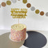 A round cake with a smooth vanilla frosting and colorful sprinkles on the sides sits on a silver cake board. Topped with a glittery gold cardstock cake topper that reads "HAPPY 80TH Birthday GRANDMA." In the background, there is a decorative garland made of circles in gold, silver, and brown. The table is covered with a mix of light and dark colored surfaces.