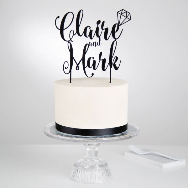 A round, white frosted cake sits on a clear glass cake stand. The cake features a black ribbon around its base. Atop the cake, there is a personalized acrylic topper in black, featuring the names "Claire" and "Mark" in decorative cursive font, along with a small diamond graphic. The background is simple and light, emphasizing the cake and topper.