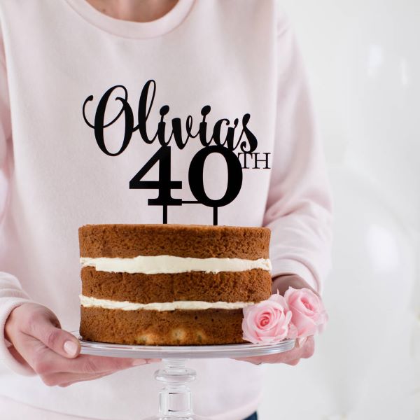 A person with a light pink sweater is holding a three-layer cake on a clear cake stand. The cake is undecorated except for two layers of cream filling and is topped with a black acrylic cake topper that reads "Olivia&
