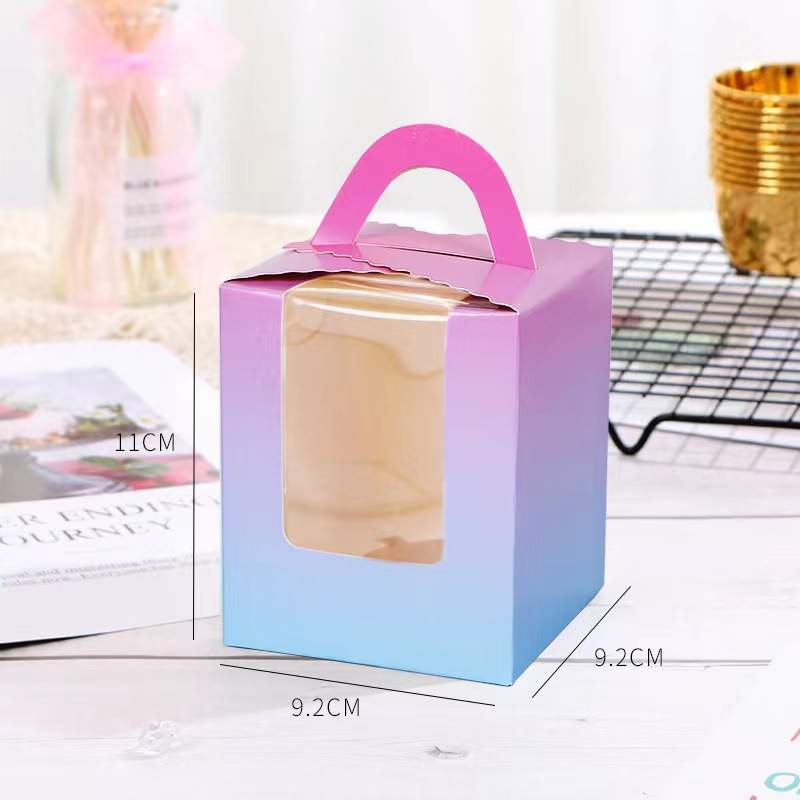 A small cupcake box designed for single portions, featuring a pink handle and a transparent window on the front. The box has a gradient color design, transitioning from pink to blue. Dimensions indicated on the box include a height of 11 cm and a width of 9.2 cm. It is placed on a table with other items visible, including a magazine and a wire cooling rack.