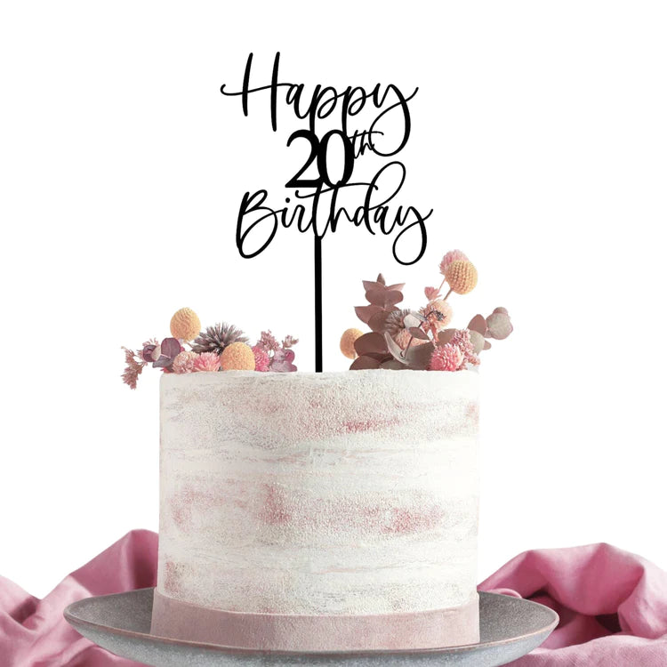 A round cake with a textured white and light pink frosting sits on a silver cake stand. Topped with a black acrylic cake topper reading "Happy 20th Birthday," the cake is also adorned with colorful dried flowers and foliage, giving it a festive look. A soft pink fabric drapes beside the cake.