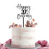 A round cake with a textured white and light pink frosting sits on a silver cake stand. Topped with a black acrylic cake topper reading "Happy 20th Birthday," the cake is also adorned with colorful dried flowers and foliage, giving it a festive look. A soft pink fabric drapes beside the cake.