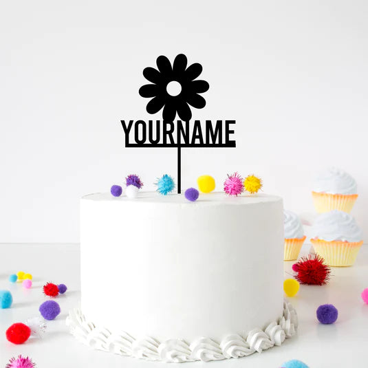 A white frosted cake with a flower-shaped acrylic topper that features the text "YOURNAME" below the flower. The cake is decorated with colorful pom-poms and is surrounded by pastel cupcakes in the background.