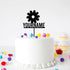 A white frosted cake with a flower-shaped acrylic topper that features the text "YOURNAME" below the flower. The cake is decorated with colorful pom-poms and is surrounded by pastel cupcakes in the background.
