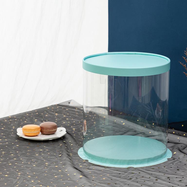 A round PVC tall cake box with a turquoise lid sits on a dark table covered with a starry tablecloth. Next to the cake box, there is a white plate holding two macarons?one orange and one chocolate?showing a dessert display. The background features a blue wall and soft, draped fabric.