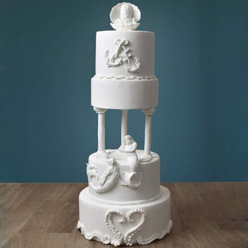 The image features a three-tiered white cake designed with intricate decorations. The top tier is adorned with a shell motif, while the middle tier displays a chain design. The lower tier is supported by four decorative pillars and features sculpted figures, including a mermaid and a dragon. The base of the cake is embellished with a heart shape made of pearls. The cake is set against a teal background, highlighting its elegant design.