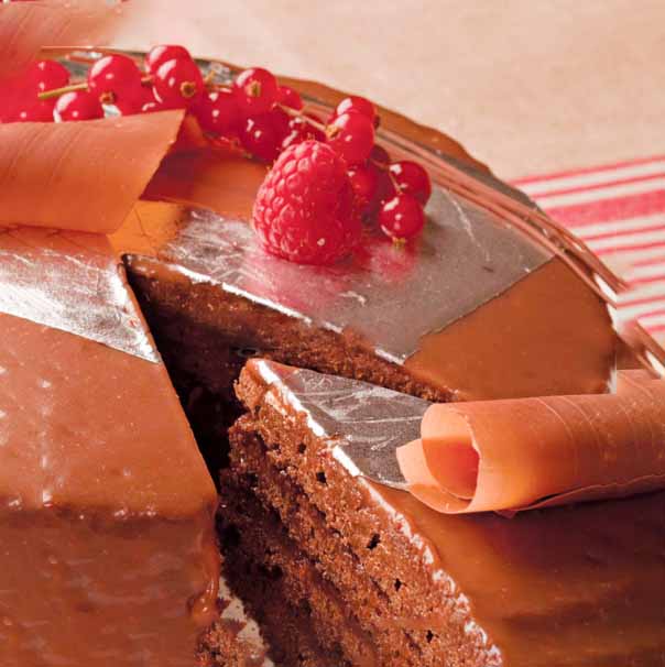 A close-up image of a chocolate cake, partially sliced to reveal its layers. The top is decorated with shiny edible silver leaves and pieces of chocolate curls. Fresh raspberries and red currants are placed on top for garnish. The background shows a light-colored surface with a subtle striped pattern.
