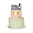 A two-tier birthday cake featuring a pink top tier with a gold heart design, and a bottom tier decorated with white ruffles and gold accents. Atop the cake is a black custom cake topper that reads "Happy Birthday" with a silhouette of a soccer player in action. The cake is presented on a circular white plate against a white background.