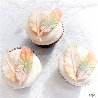 Three cupcakes are displayed on a shiny surface. Each cupcake has a smooth white frosting topped with an intricately designed multi-colored feather made from silicone. The feathers feature soft pastel hues, including shades of pink, green, and yellow, resembling realistic feathers. The cupcakes have dark brown liners, contrasting with the light frosting and vibrant feathers.
