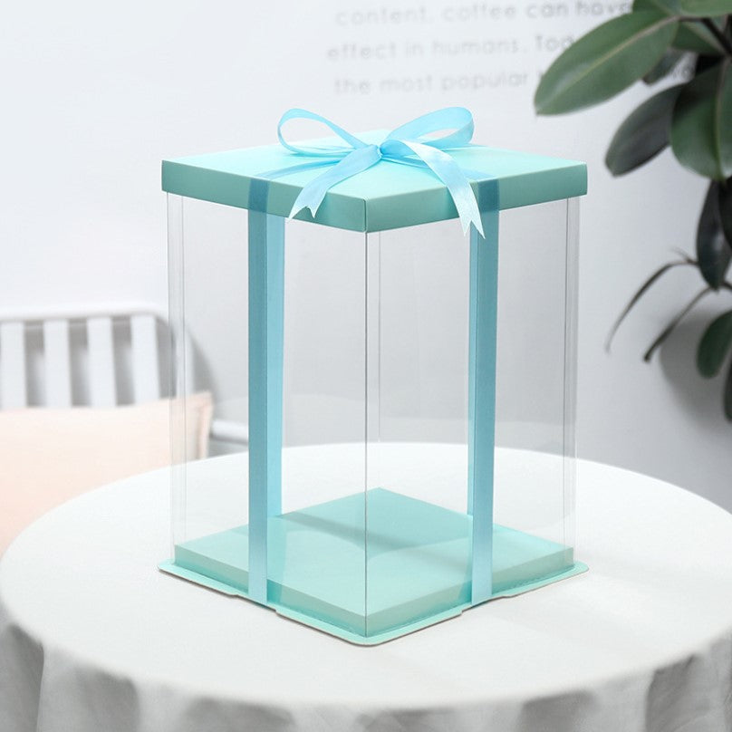 A square cake box, measuring 14 x 14 x 14 inches, featuring a light blue top and matching ribbon. The box has transparent sides, allowing visibility of the contents inside. It is placed on a round table with a light-colored tablecloth, and there are subtle decorative elements in the background, including a green plant and possibly furniture. The overall aesthetic is clean and modern.