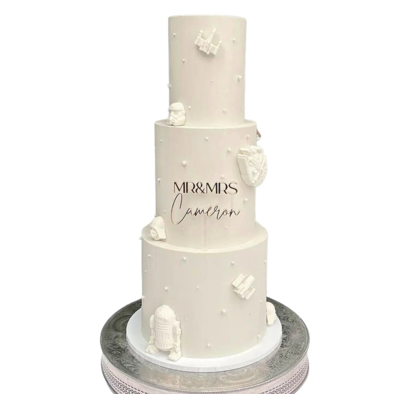 A three-tiered white wedding cake with a smooth finish. The middle tier features the text "MR & MRS" followed by the name "Cameron" in elegant script. The cake is adorned with small white spheres and decorative elements resembling popular sci-fi characters and symbols, such as a stormtrooper helmet and a droid. The cake is placed on a decorative silver cake stand.