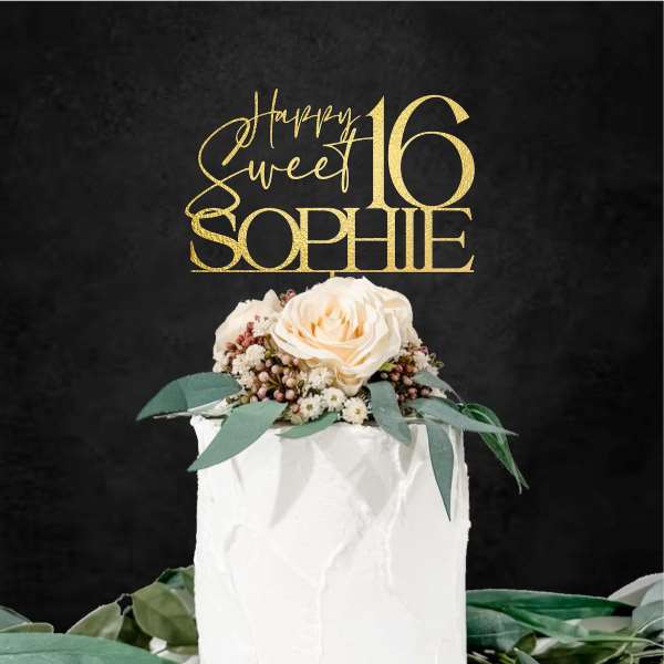An elegant white cake topped with a floral arrangement featuring a light pink rose and greenery. Above the cake, there is a customized acrylic topper that reads "Happy Sweet 16 SOPHIE" in gold lettering against a dark background. The overall presentation is festive and suitable for a sweet sixteen celebration.