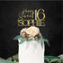 An elegant white cake topped with a floral arrangement featuring a light pink rose and greenery. Above the cake, there is a customized acrylic topper that reads "Happy Sweet 16 SOPHIE" in gold lettering against a dark background. The overall presentation is festive and suitable for a sweet sixteen celebration.
