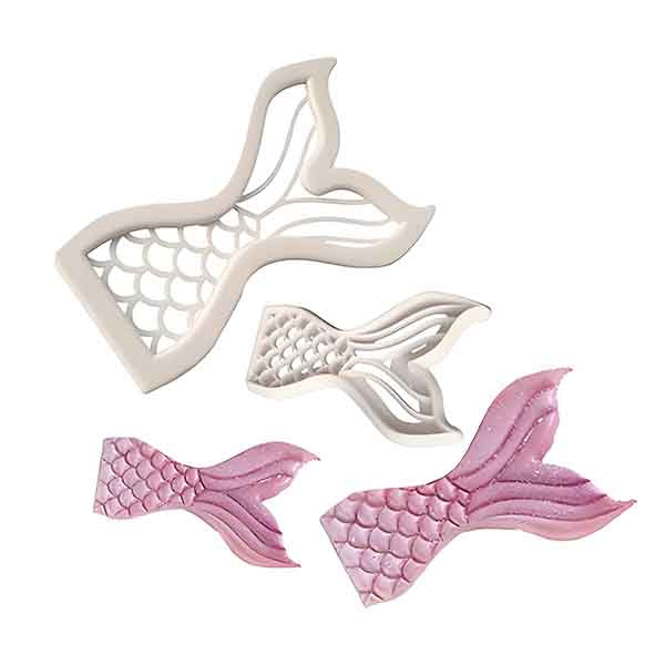Image shows a set of cookie cutters designed in the shape of mermaid tails. There are three cookie cutters: one larger, detailed cutter with a scalloped texture and two smaller cutters, one with slightly simpler details. The smaller cutters are shown in light pink, while the larger one is a white plastic. The overall design emphasizes the curves and fins associated with mermaid tails.