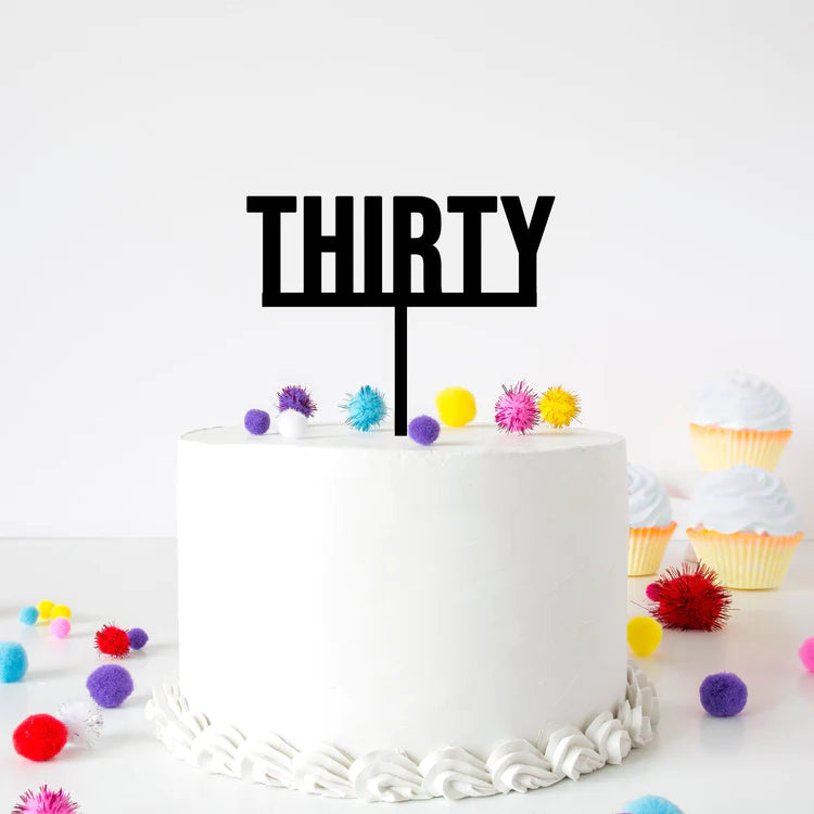 A white frosted round cake with a black acrylic cake topper that reads "THIRTY." The cake is adorned with colorful pom-poms in various sizes, including purple, yellow, red, and blue, scattered around the base. In the background, there are decorated cupcakes with white frosting and yellow wrappers. The setting is bright and festive.