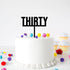 A white frosted round cake with a black acrylic cake topper that reads "THIRTY." The cake is adorned with colorful pom-poms in various sizes, including purple, yellow, red, and blue, scattered around the base. In the background, there are decorated cupcakes with white frosting and yellow wrappers. The setting is bright and festive.