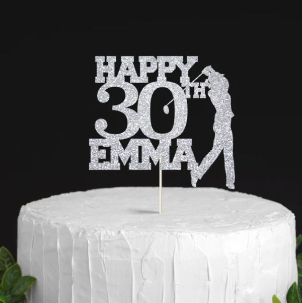 A silver glittery cake topper featuring the text "HAPPY 30th EMMA" along with a silhouette of a golfer in action, displayed on a white frosted cake with a textured surface. The backdrop is dark, highlighting the cake and topper.