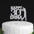 A silver glittery cake topper featuring the text "HAPPY 30th EMMA" along with a silhouette of a golfer in action, displayed on a white frosted cake with a textured surface. The backdrop is dark, highlighting the cake and topper.