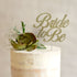 A cream-colored, frosted cake topped with a gold glitter cardstock cake topper that reads "Bride to Be." The topper is decorated with a succulent and green leaves, along with small white flowers. The background is softly blurred.