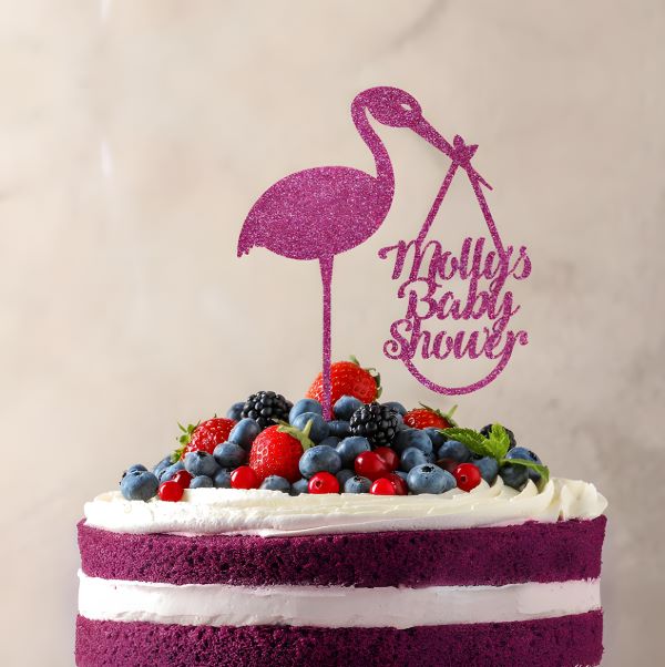 A layered cake with purple layers and white frosting topped with a variety of fresh berries, including strawberries, blueberries, and blackberries. Atop the cake is a pink glittery cake topper in the shape of a stork holding a bag, accompanied by the text "Molly&