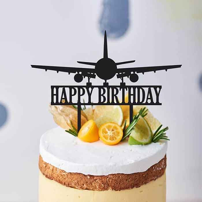 The image features a cake with a creamy white frosting and a brown crust. Topping the cake is a black acrylic cake topper shaped like an airplane, with the words "HAPPY BIRTHDAY" displayed below it. The cake is adorned with slices of lemon, lime, and orange, along with some sprigs of rosemary, adding a fresh, decorative touch. The background includes a subtle, light-colored design that enhances the focus on the cake and topper.