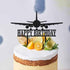 The image features a cake with a creamy white frosting and a brown crust. Topping the cake is a black acrylic cake topper shaped like an airplane, with the words "HAPPY BIRTHDAY" displayed below it. The cake is adorned with slices of lemon, lime, and orange, along with some sprigs of rosemary, adding a fresh, decorative touch. The background includes a subtle, light-colored design that enhances the focus on the cake and topper.